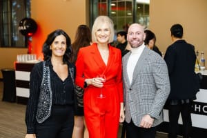 Three people posing together at an indoor event, one holding a wine glass. Dressed in formal attire, they capture the essence of event photography amid a room full of elegantly dressed attendees.