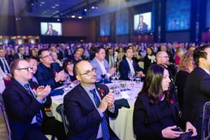 A large audience seated at round tables in a dimly lit venue watches a presentation captured through event videography. Some are clapping.