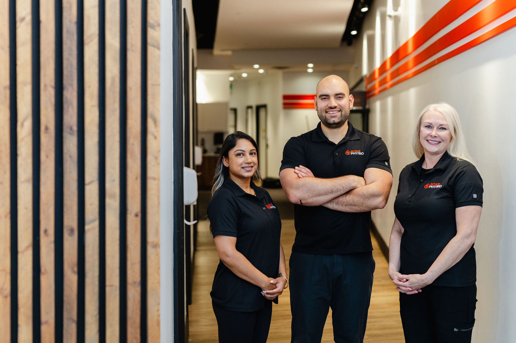 Three people stand in a modern hallway with wooden walls and red accents, each wearing matching black polo shirts adorned with a logo. This setting provides an ideal backdrop for professional headshots, capturing their polished and unified appearance.
