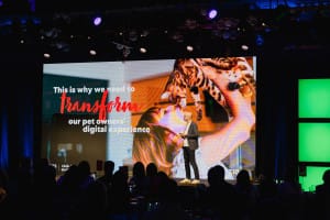 A person presents on stage, as an event photographer captures the moment a large screen displays a message about transforming pet owners' digital experience, accompanied by an image of someone holding a cat.