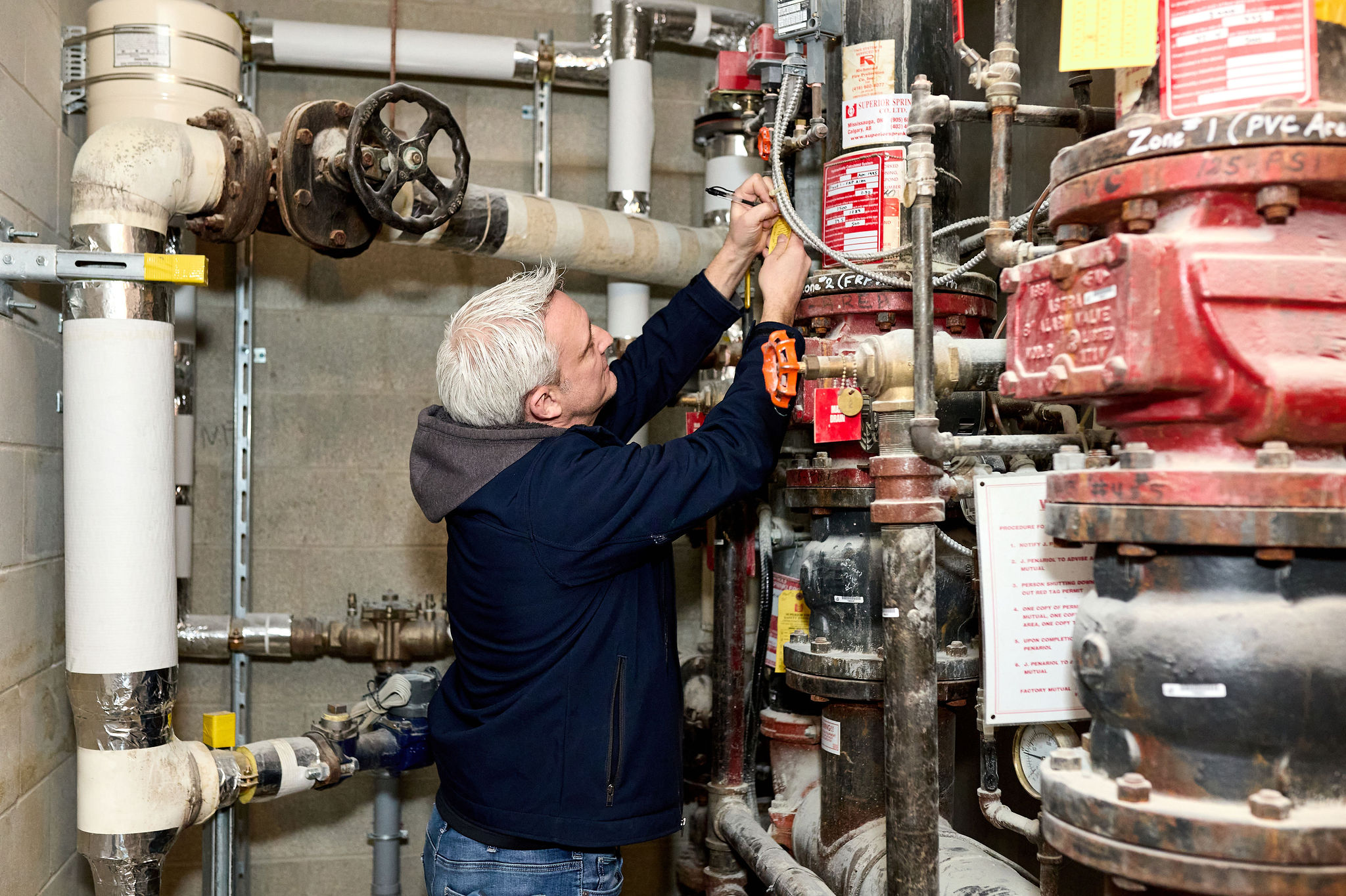 A person with gray hair is adjusting a valve in a room filled with large pipes and mechanical equipment, reminiscent of the behind-the-scenes intricacies often captured in event videography.