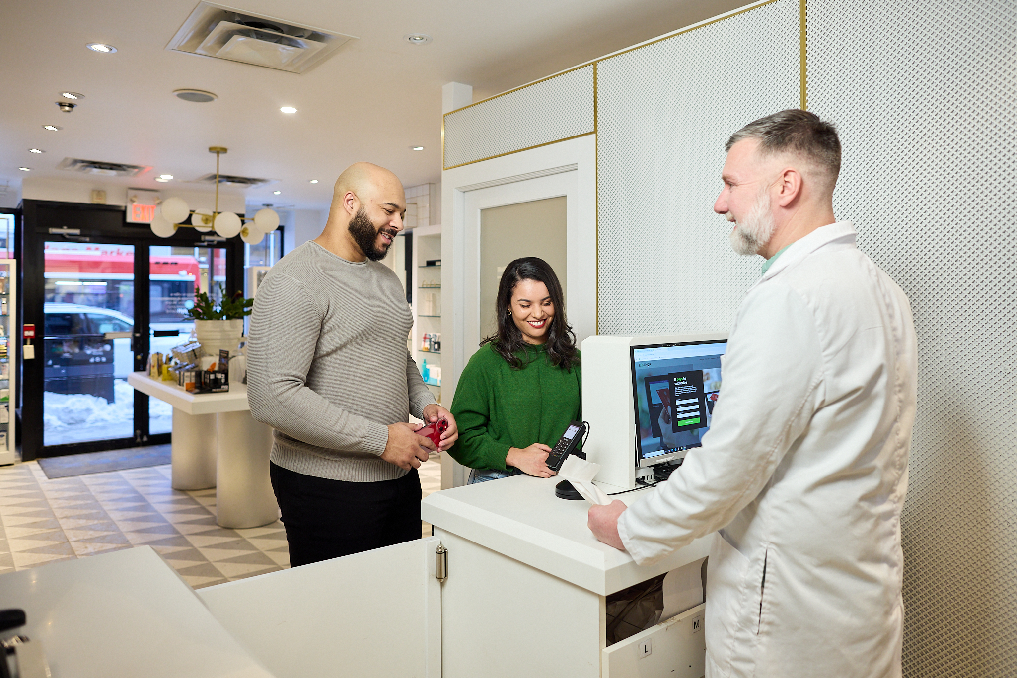A man and woman stand at a pharmacy counter, interacting with a pharmacist in a white coat; the woman holds a phone near a payment terminal, capturing a moment of corporate lifestyle photography.