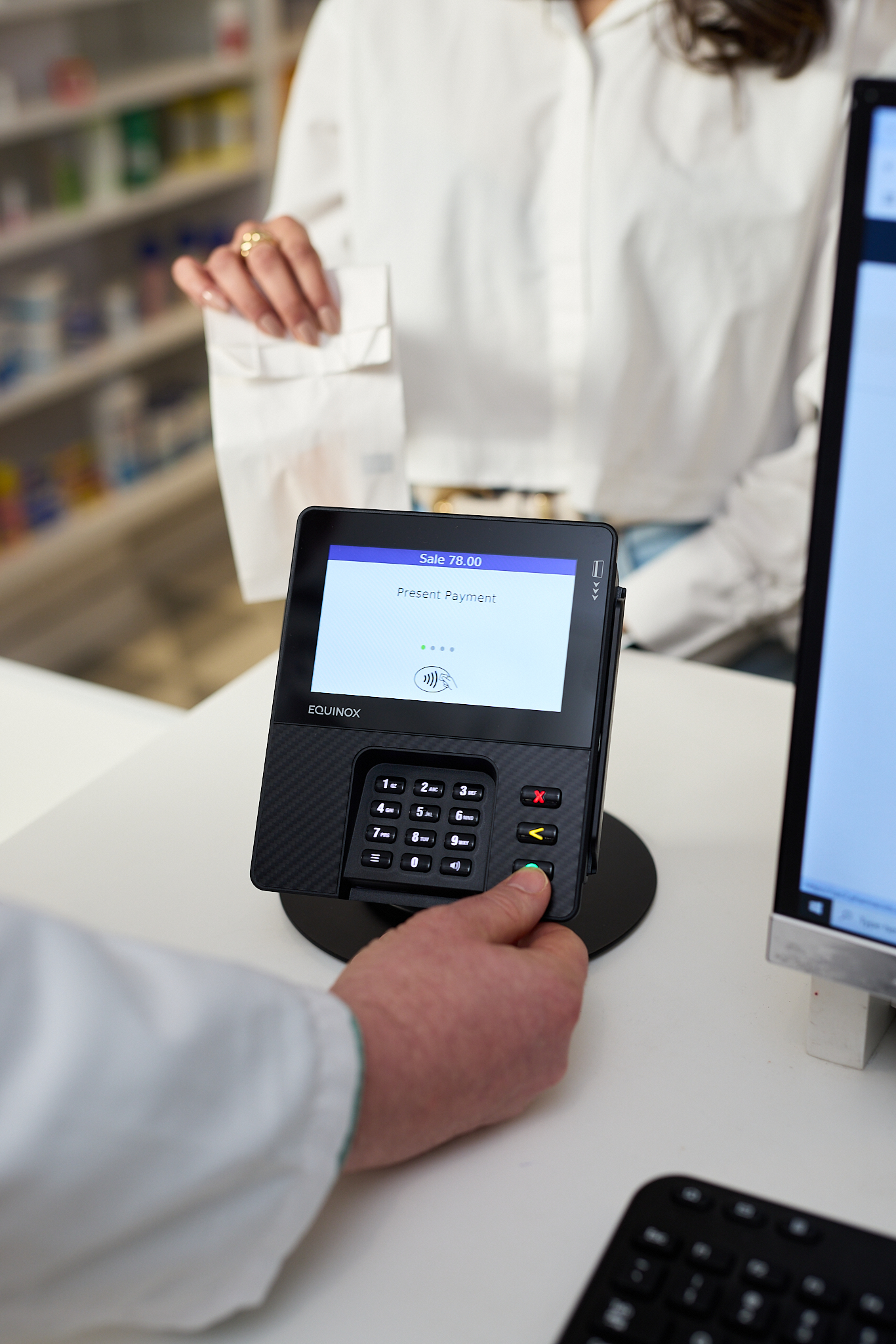A person in a white coat holds a payment terminal while another, holding a paper bag, prepares to pay at a pharmacy counter—capturing the essence of corporate lifestyle photography in a healthcare setting.