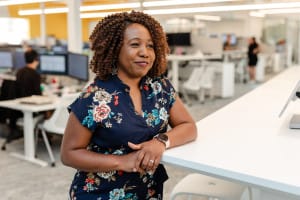 A woman in a floral dress leans on a white table in a modern, open-plan office environment.