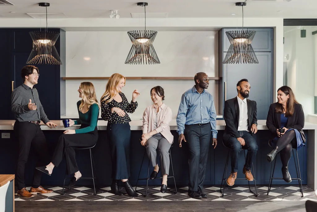 Seven professionally dressed people sit and stand around a modern office kitchen counter, conversing and smiling under hanging light fixtures.