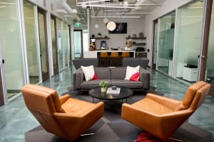 Modern office lounge with two brown leather chairs, a gray sofa, a round coffee table, and a kitchenette in the background. Glass-walled offices line both sides.