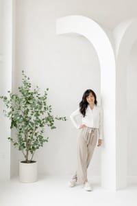 A woman stands indoors leaning against a white arch, wearing a white blouse and beige pants. A potted plant is positioned to her left. The room has a minimalist, light-colored design.