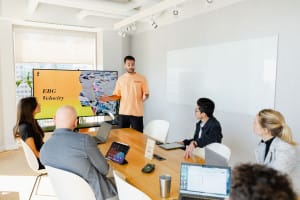 A man stands and presents a slide titled "EBG Velocity" to four colleagues seated around a conference table with laptops and tablets.