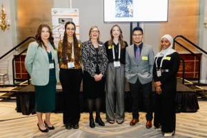 Six people stand in a row smiling, dressed in business attire, exemplifying the perfect moment in business photography. They pose confidently in front of a presentation screen and stage steps.