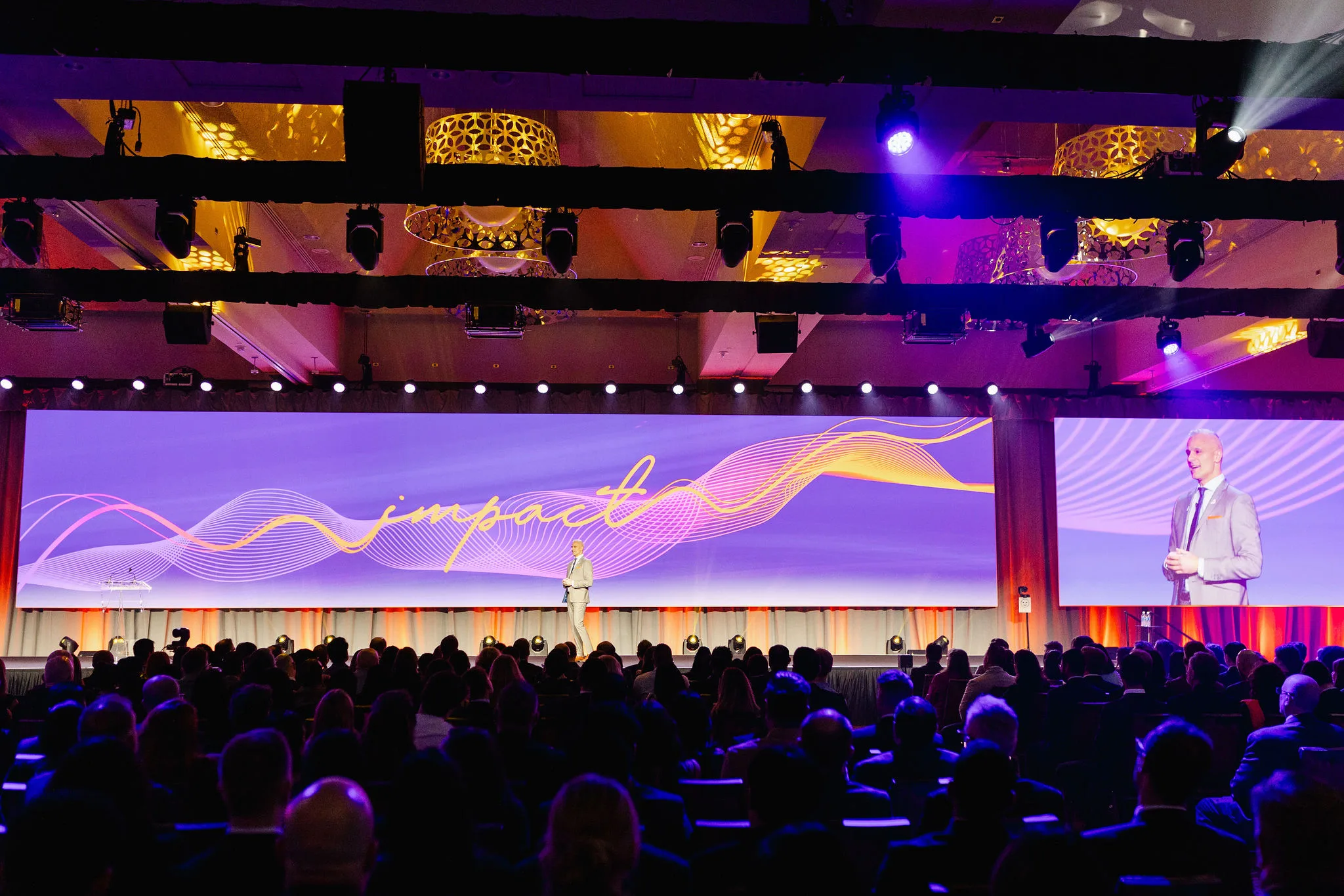 A speaker stands onstage in front of an audience, with the word "impact" and colorful wave graphics displayed on a large screen behind him, captured perfectly as part of Grand Opening Photography.