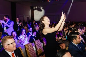 A woman stands to capture a Grand Opening Photography moment on her phone in a crowded, seated audience at an indoor event, while others clap and watch the stage.
