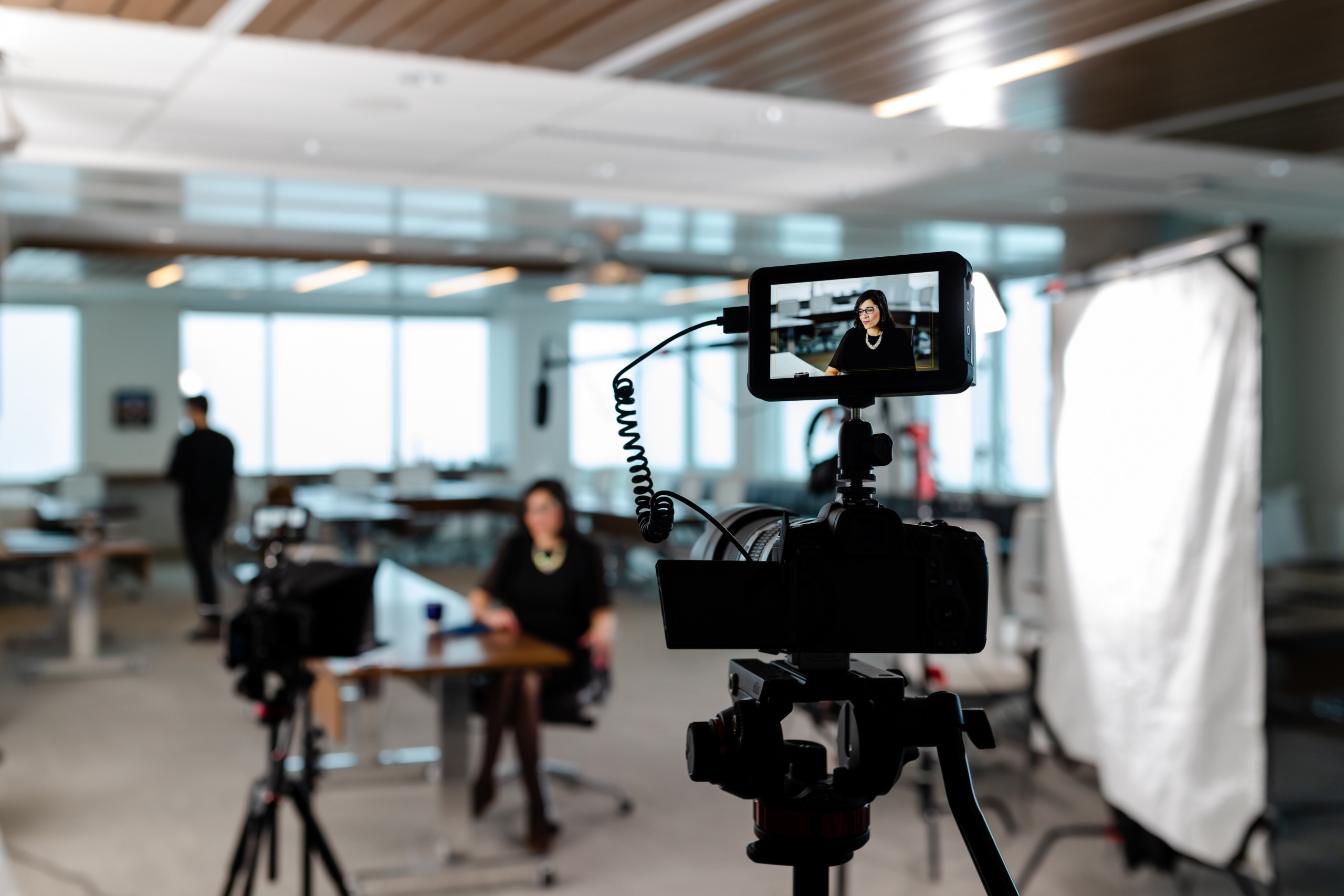 A camera records a person seated at a table in a modern office studio, with lighting equipment and another person standing in the background.