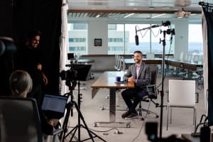 A man in business attire sits at a table in an office, being filmed by a crew with cameras, lights, and microphones for what appears to be an interview or video shoot.