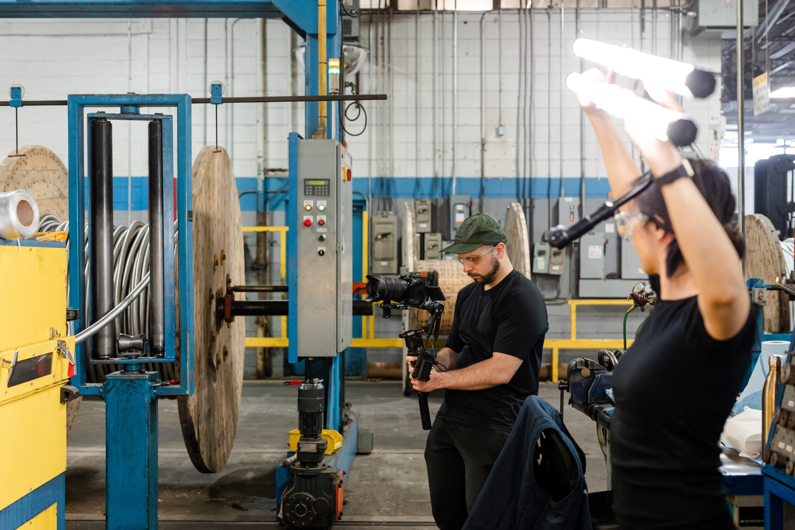 Two people work in an industrial setting; one operates a camera near large cable spools, while another holds up lights in the foreground.