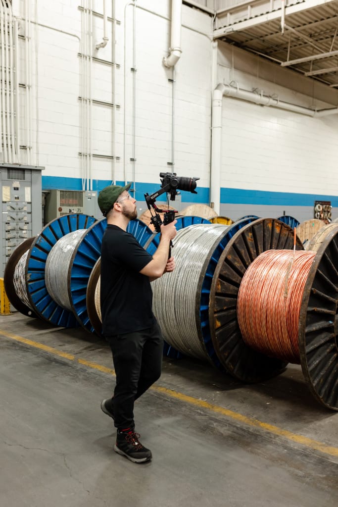 A man wearing black clothing and a green cap films with a camera stabilizer in an industrial setting with large spools of cable in the background.