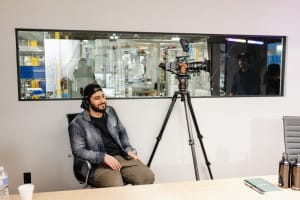 A man sits in a chair near a professional camera on a tripod in an office; a window behind reveals a lab or factory area with people and equipment.