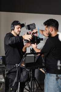 Two men adjust a professional video camera mounted on a tripod in an indoor setting with various film equipment visible.