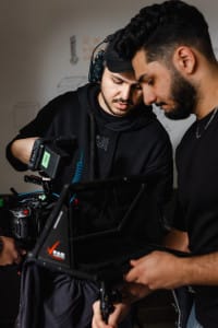 Two men operate video production equipment, one adjusting a camera and the other holding a teleprompter, with a whiteboard in the background.
