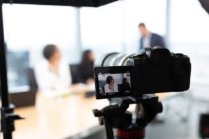 A digital camera on a tripod records a business meeting with three people sitting at a conference table in a modern office.