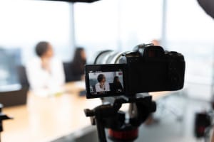 A digital camera records two people seated at a table in an office, with the camera’s display screen showing the scene in focus.