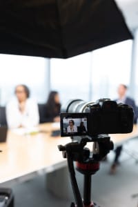 A digital camera on a tripod records a business meeting with three people seated at a conference table in a bright office.