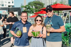 Three people standing outdoors, smiling and holding tropical drinks served in fruit, perfect for a vibrant corporate photography session. Red umbrellas and other people are visible in the background on a sunny day.