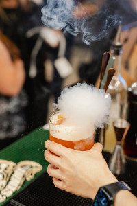 A hand holds a reddish cocktail emitting smoke or vapor at a bar, with bottles and bar tools visible in the background.