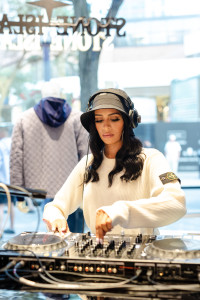 A woman wearing headphones and a white sweater is DJing at a mixing console indoors, with clothing visible in the background.