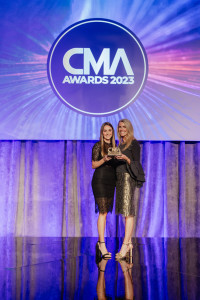 Two women stand on stage holding an award at the CMA Awards 2023, with the event logo displayed on a screen behind them.