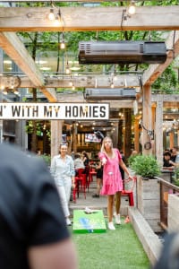 Two women playing cornhole in an outdoor area with string lights and wooden structures, while people sit at tables in the background.
