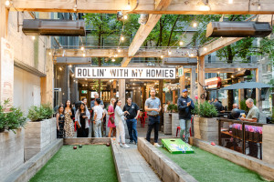 A group of people stand and play games in an outdoor urban patio with string lights and a sign that reads "ROLLIN' WITH MY HOMIES.