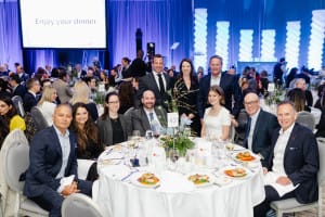 A group of people in formal attire sit and stand around a table set for dinner at a large, elegant event. This moment of celebration and connection is perfectly captured through professional event photography.