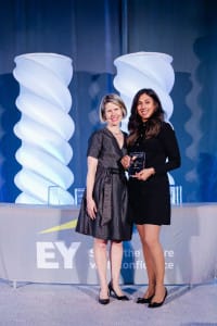 Two women stand in front of an EY-branded table at an event, one holding a glass award. Professionally dressed and smiling at the camera, they capture the spirit of event photography.