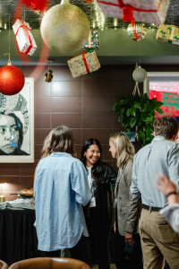 A group of people stand and talk at a festive indoor gathering, with hanging ornaments and wrapped gifts decorating the ceiling.
