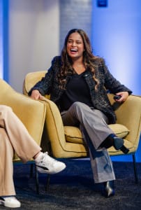 A woman sitting on a yellow chair, smiling and laughing, dressed in a dark blazer, black top, gray pants, and black shoes, with another person partially visible beside her.