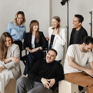A group of seven people, casually dressed, sit and stand together indoors, smiling and laughing in a relaxed, friendly atmosphere—capturing the candid side of corporate photography.