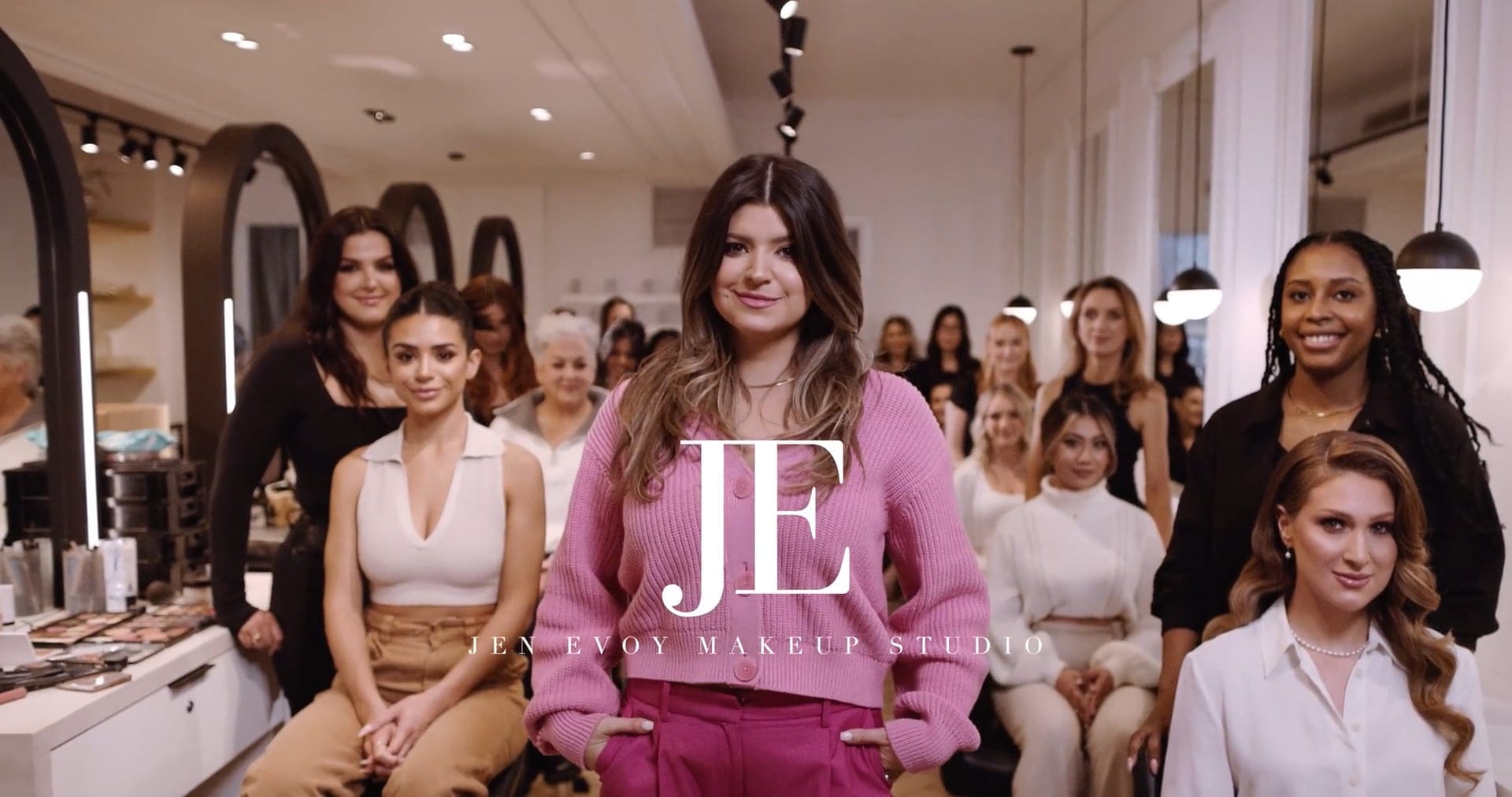 A creative team of women pose together inside a makeup studio, with the studio's logo "JE" and "Jen Envoy Makeup Studio" displayed across the center.