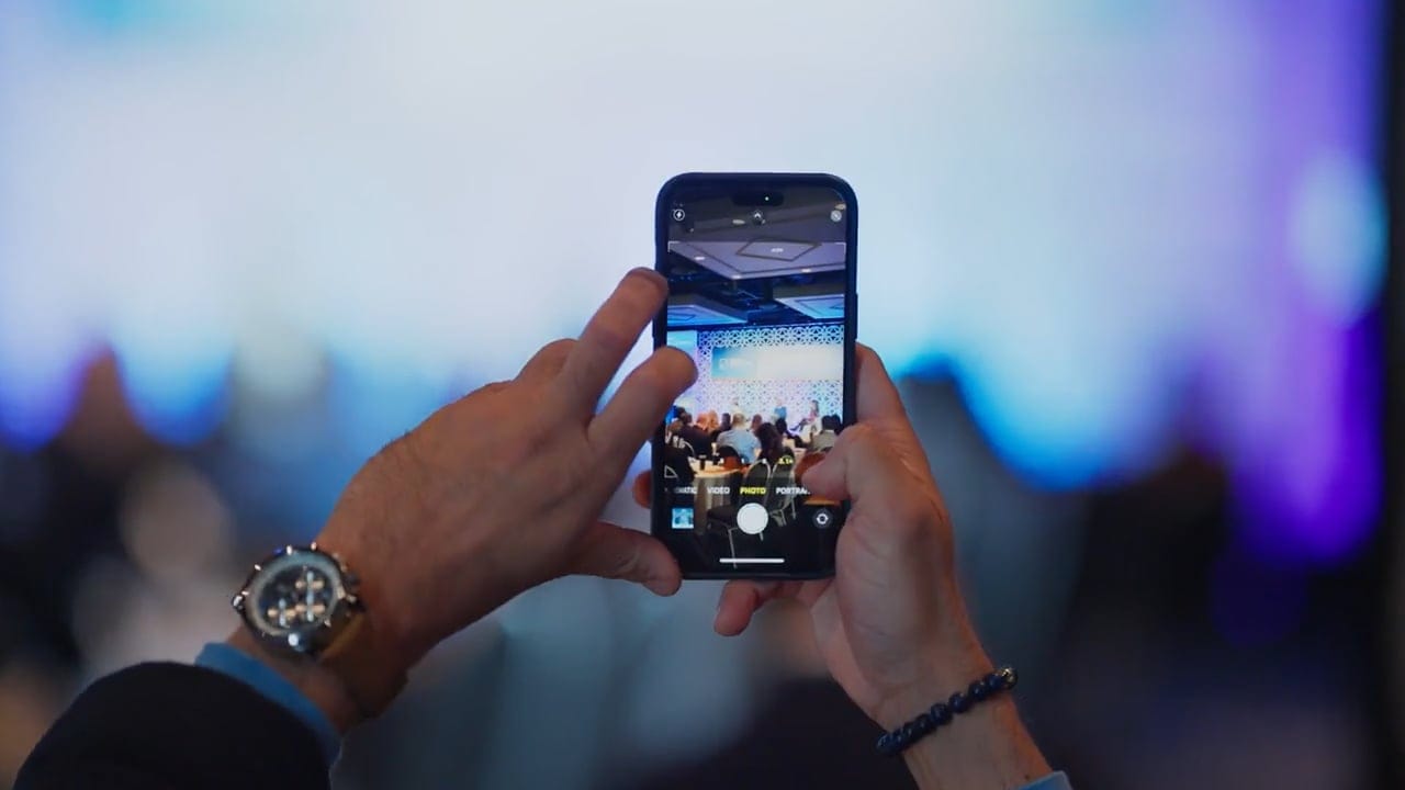 A person holds a smartphone, snapping a photo of a blurred event or conference where the creative team and other attendees are visible on stage through the phone screen.