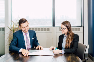 Two people in business attire sit at a round table in an office, looking at documents together. Large windows and a plant are visible in the background.