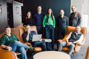 Eight people pose for a group photo in an office lounge area with modern chairs; one person holds a laptop.