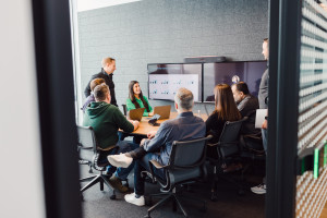 A group of people sit around a conference table with laptops, viewing charts on a large screen, during a business meeting in a modern office.