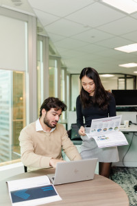 A man sits at a desk working on a laptop while a woman stands beside him holding and discussing a printed report in a modern office setting.