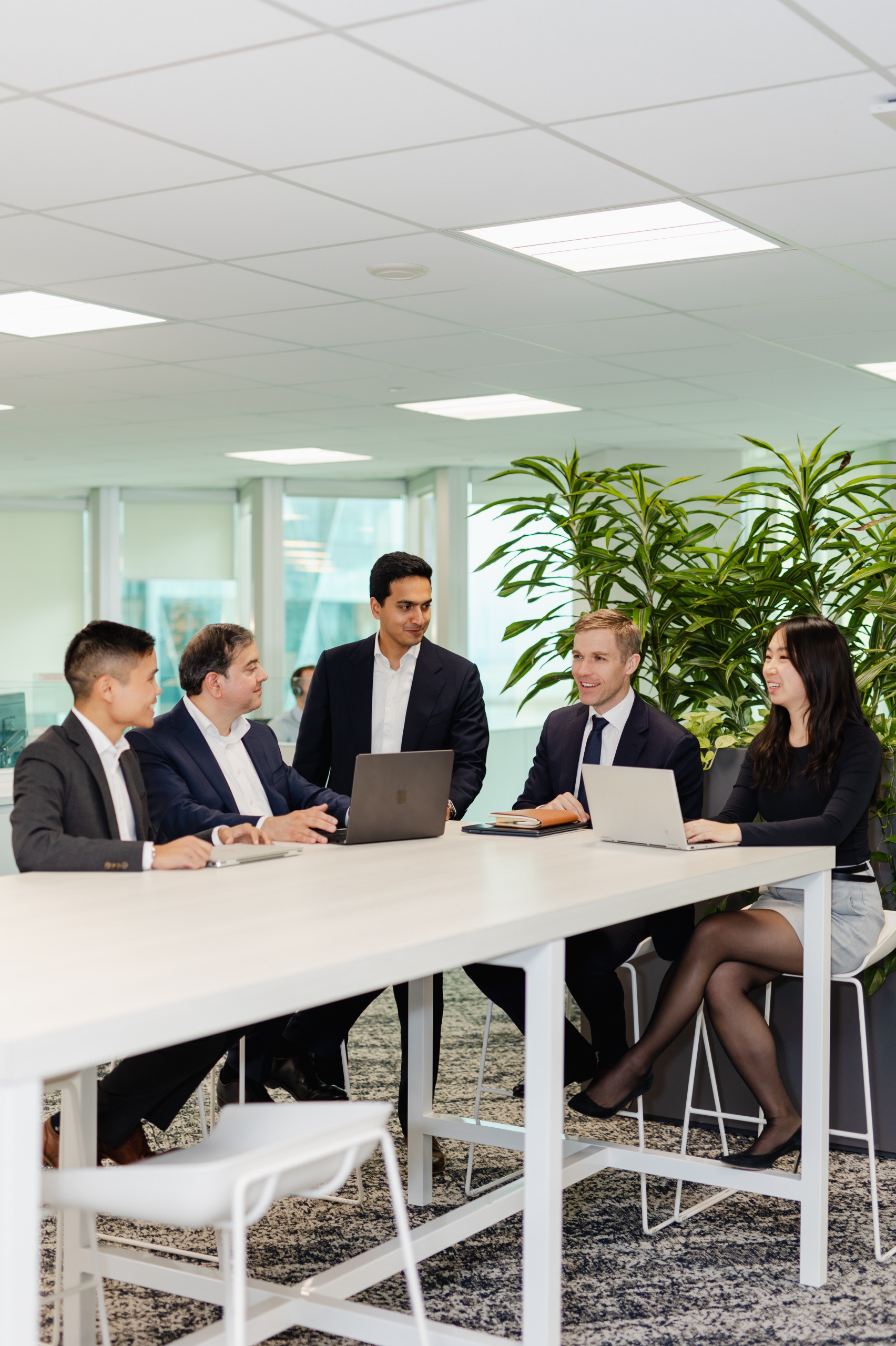 Five people in business attire sit and stand around a meeting table with laptops, having a discussion in a modern office space with large windows and green plants.