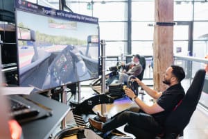 A man operates a racing simulator in a modern room with large windows, captured in dynamic event photography, while another person uses a similar setup in the background.