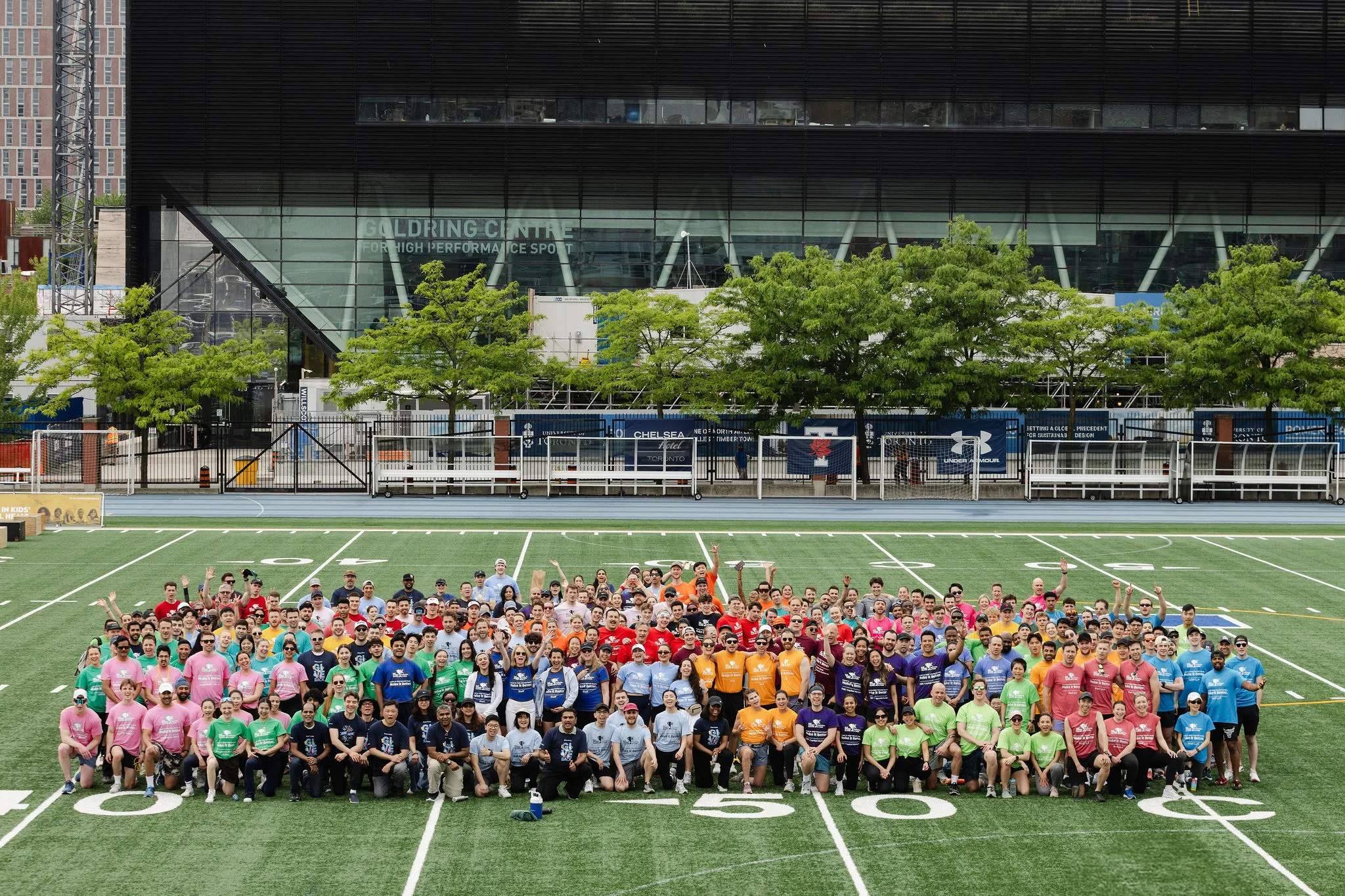 A large group of people in colorful shirts pose for an event photography session on a football field in front of a modern building.