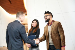 Three people in business attire greet each other; two men shake hands while a woman stands between them, smiling, in a modern office setting.
