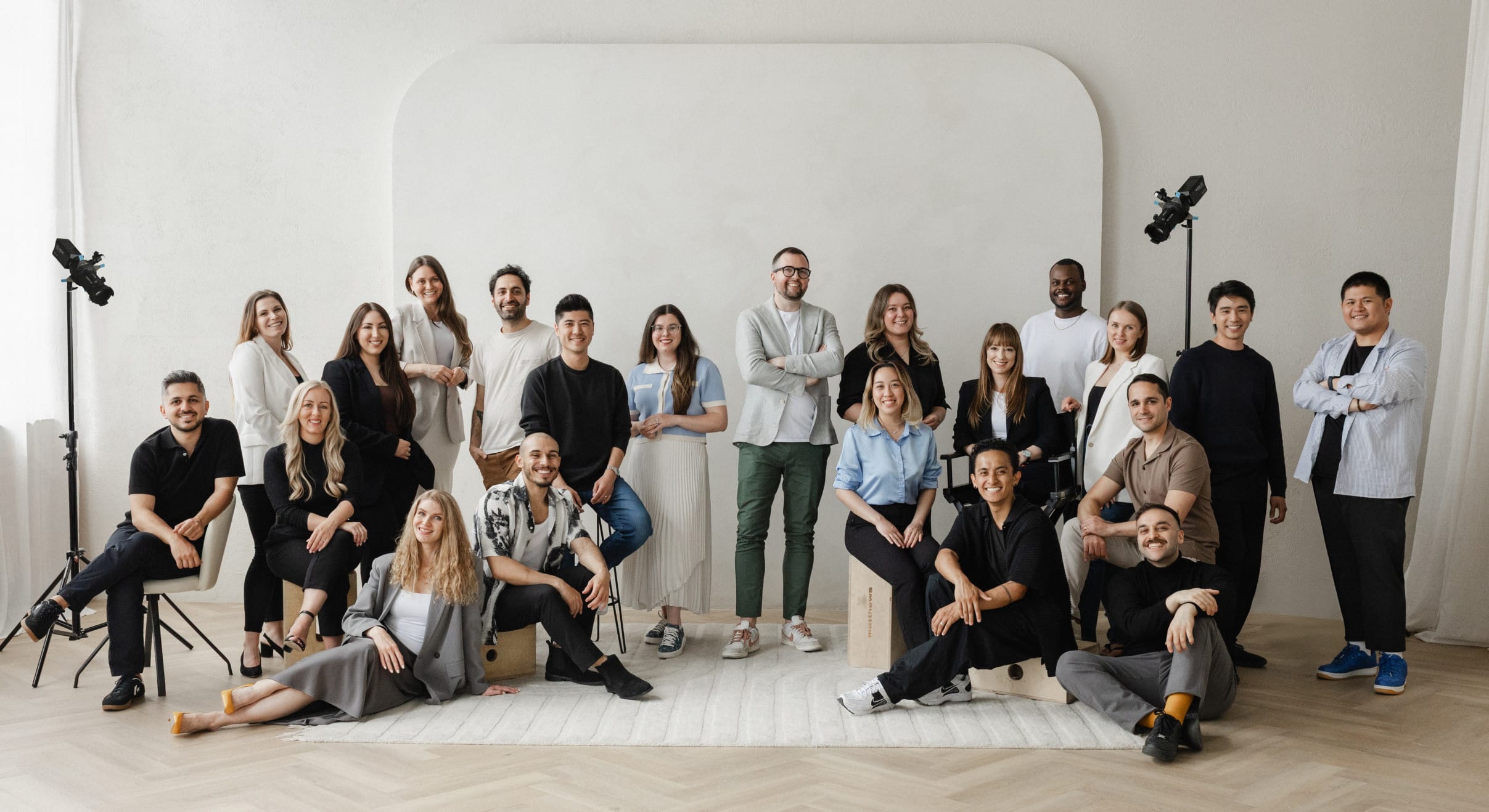 A diverse creative team of 20 people poses together in a bright studio space, standing and sitting in front of a neutral backdrop with two studio lights visible.