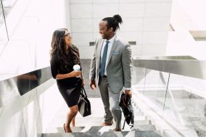 Two professionally dressed people walk up a glass staircase, holding a briefcase and coffee, and smiling at each other in a modern office building.