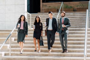 Four professionals in business attire walk down outdoor steps, each holding coffee cups, appearing to converse and smile.