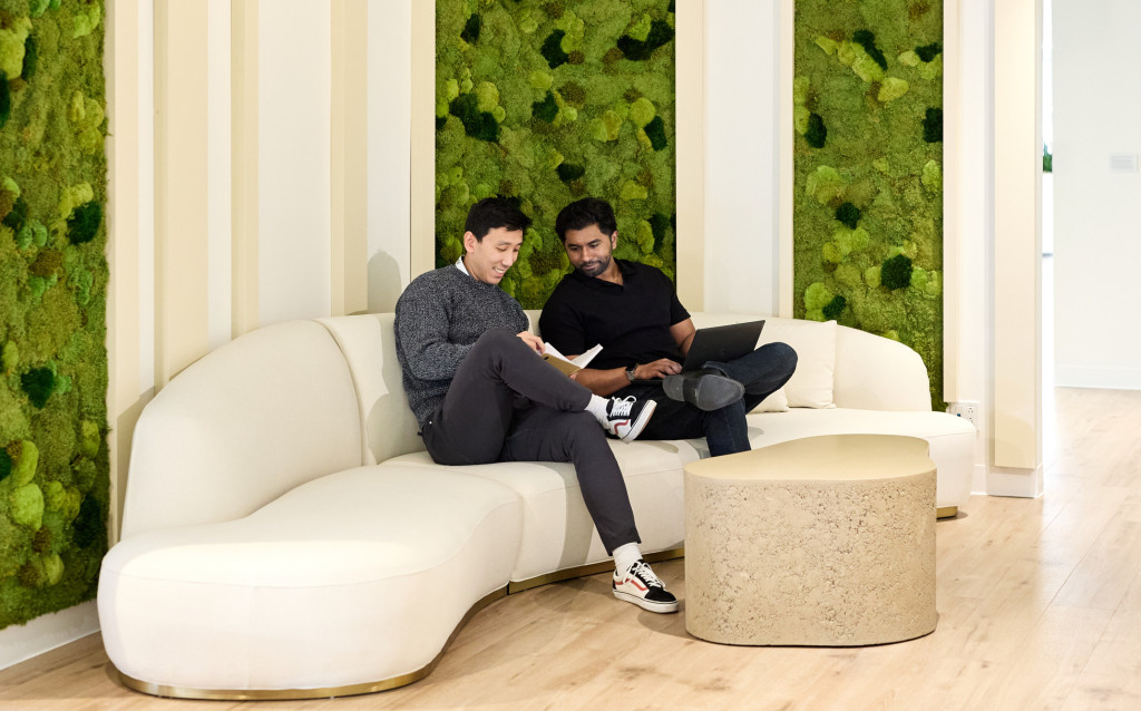 Two people sit on a curved white couch in a modern office lounge with green plant walls, reading and working together at a round table.